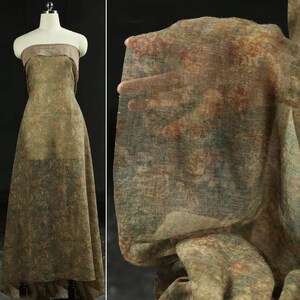 May include: A sheer, patterned fabric in shades of olive green, brown, and red is draped over a dress form. The fabric has a delicate, almost transparent quality, with a subtle floral or abstract design. The dress form is a neutral color, allowing the fabric's pattern to stand out.