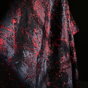 May include: A close-up of a fabric with a dark background. The fabric is primarily black with a textured pattern and is accented with vibrant red metallic flecks and streaks, creating a shimmering effect.