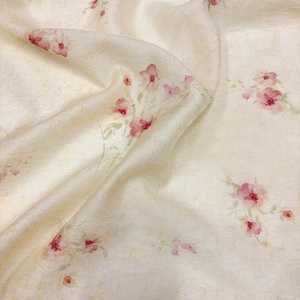 May include: A piece of off-white fabric with a textured surface featuring a scattered pattern of small, delicate pink flowers and thin green stems. The material is draped in soft folds, highlighting the subtle variations in the light cream colour.