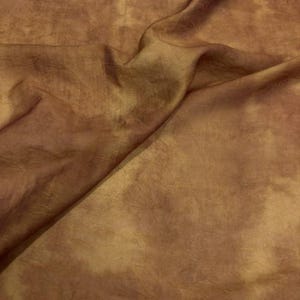 May include: Close-up of a folded fabric with a mottled appearance. The fabric has a warm color palette, with shades of brown, gold, and hints of reddish-brown. The material appears to be soft and flowing, with subtle textures and folds.