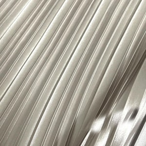 Puede incluir: Primer plano de una tela con pliegues verticales. La tela tiene un color gris plateado nacarado, con la luz reflejándose en los pliegues. Los pliegues corren diagonalmente, creando una superficie texturizada.