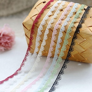 Puede incluir: Una selección de delicadas cintas de encaje en varios colores, incluyendo burdeos, blanco, rosa, verde claro y negro. Las cintas tienen un borde festoneado y están dispuestas en diagonal, con una cesta de mimbre en el fondo.