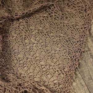 Pode incluir: Close-up de um tecido castanho de trama aberta com um padrão delicado, semelhante a uma rede. O tecido está drapeado sobre uma superfície de madeira, mostrando sua textura e design. O material parece macio e flexível.