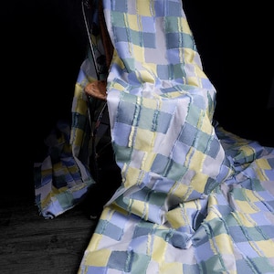 May include: A draped fabric with a patchwork design in shades of blue, green, and yellow. The fabric is draped over a wooden chair, showcasing its texture and pattern. The squares are approximately 4 inches by 4 inches.