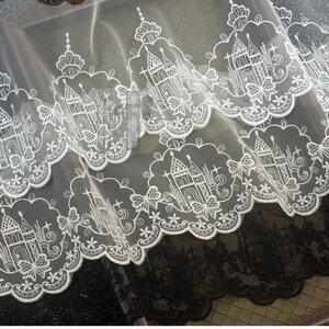 Peut inclure: Un tissu en dentelle blanche pr&eacute;sentant un motif r&eacute;p&eacute;titif de ch&acirc;teaux et de papillons brod&eacute;s avec un bord festonn&eacute;. Le travail complexe du fil blanc est pos&eacute; sur un fond en maille fine et transparente, cr&eacute;ant un design textile d&eacute;taill&eacute;.