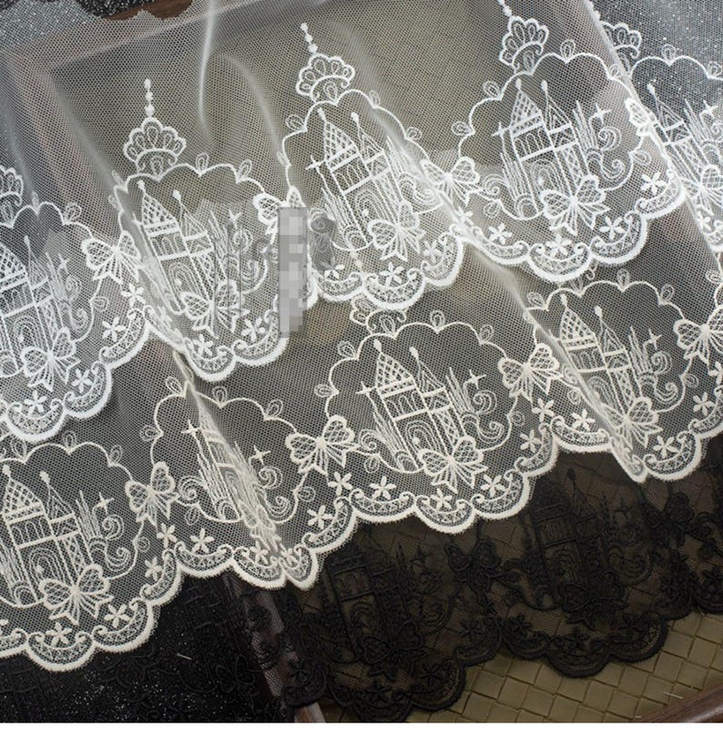 Peut inclure: Un tissu en dentelle blanche pr&eacute;sentant un motif r&eacute;p&eacute;titif de ch&acirc;teaux et de papillons brod&eacute;s avec un bord festonn&eacute;. Le travail complexe du fil blanc est pos&eacute; sur un fond en maille fine et transparente, cr&eacute;ant un design textile d&eacute;taill&eacute;.