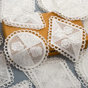 May include: White lace doilies in various shapes, including round, square, and diamond. Each doily features intricate floral patterns and scalloped edges. The doilies are arranged on a wooden surface, showcasing their delicate details.