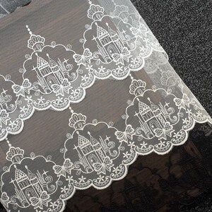 Peut inclure: Un tissu en dentelle blanche pr&eacute;sentant un motif r&eacute;p&eacute;titif de ch&acirc;teaux complexes entour&eacute;s de bordures festonn&eacute;es. Le design inclut des accents floraux d&eacute;licats, des formes d'&eacute;toiles et des d&eacute;tails rappelant des papillons en fil blanc.