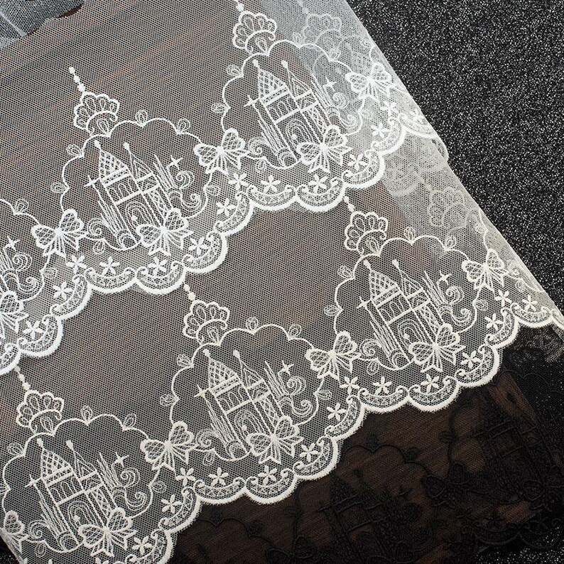 Peut inclure: Un tissu en dentelle blanche pr&eacute;sentant un motif r&eacute;p&eacute;titif de ch&acirc;teaux complexes entour&eacute;s de bordures festonn&eacute;es. Le design inclut des accents floraux d&eacute;licats, des formes d'&eacute;toiles et des d&eacute;tails rappelant des papillons en fil blanc.