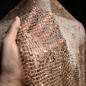 Könnte beinhalten: Ein transparenter Stoff mit offenem Strickmuster aus metallischem, bronzefarbenem und kupferfarbenem Garn. Das Material hat eine lockere, gitterartige Struktur mit kleinen, unregelmäßigen weißen und schwarzen Sprenkeln, die in die Fäden eingewebt sind.