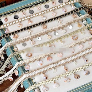 May include: Assortment of decorative trims with seashell, butterfly, and sequin embellishments. The trims feature various colors, including white, black, and gold, and are suitable for crafting and embellishing projects. The trims are displayed on a light-colored surface.