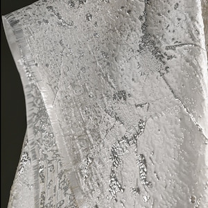 Op de afbeelding: Close-up van een getextureerde, zilverkleurige stof met een metallic glans. De stof heeft een gelaagde uitstraling met onregelmatige patronen en een subtiel, glinsterend effect. De randen zijn netjes afgesneden en onthullen een gedetailleerde weefsel.
