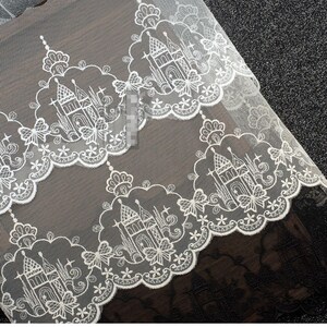 Peut inclure: Un tissu en dentelle blanche pr&eacute;sentant un motif r&eacute;p&eacute;titif de ch&acirc;teaux complexes entour&eacute;s de bordures festonn&eacute;es. Le design inclut des accents floraux d&eacute;licats et des formes d'&eacute;toiles, sur un fond en maille transparente avec une finition festonn&eacute;e.