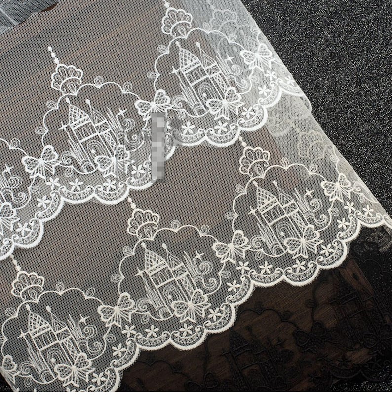 Peut inclure: Un tissu en dentelle blanche pr&eacute;sentant un motif r&eacute;p&eacute;titif de ch&acirc;teaux complexes entour&eacute;s de bordures festonn&eacute;es. Le design inclut des accents floraux d&eacute;licats et des formes d'&eacute;toiles, sur un fond en maille transparente avec une finition festonn&eacute;e.