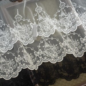 Peut inclure: Une bordure en dentelle blanche pr&eacute;sentant un motif r&eacute;p&eacute;titif de ch&acirc;teaux et de papillons brod&eacute;s avec un bord festonn&eacute;. Le design complexe est cousu sur un tissu en maille transparente, mettant en valeur des motifs architecturaux d&eacute;taill&eacute;s.