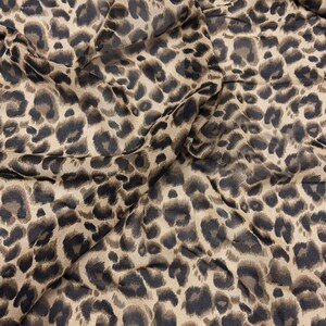 Puede incluir: Una tela ligera con un patrón de estampado de leopardo repetitivo en tonos beige, marrón claro y marrón oscuro. El material está drapeado para mostrar pliegues y arrugas, resaltando la textura suave y las formas orgánicas de las manchas.
