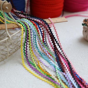 Puede incluir: Una colección de adornos de ric rac ondulados multicolores, que incluyen amarillo, verde, morado y rosa. El adorno se muestra sobre una superficie blanca, con una regla de madera y una cesta de alambre blanca en el fondo. El ric rac se utiliza para coser y hacer manualidades.