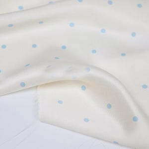 May include: Off-white fabric with a delicate sheen, scattered with small, light blue polka dots. The fabric is gently folded, revealing its texture and drape. The edge of the fabric is unfinished, showing the raw edge.