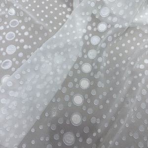 May include: White sheer fabric with a pattern of raised, textured circles of varying sizes. The fabric is draped, creating folds and shadows that add depth to the design. The circles are evenly spaced across the material.