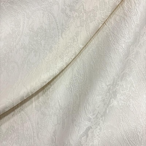 May include: Off-white fabric with a detailed paisley pattern. The fabric is folded, revealing the texture and design. The pattern is a repeating floral and curved design, creating a textured surface. The fabric appears to be a medium weight.