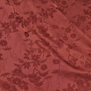May include: Close-up of a fabric with a floral pattern in shades of red. The design features various flowers and leaves, creating a textured appearance. The fabric is draped, showing folds and shadows that add depth to the design.