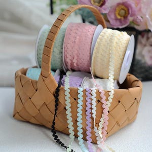 Peut inclure: Un panier en osier rempli de bobines de ruban décoratif coloré. Les bobines sont dans des tons pastel de rose, vert et jaune. Plusieurs brins de ruban à bords ondulés de différentes couleurs, dont noir, vert menthe, lavande et pêche, sortent du panier.
