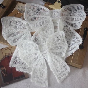 May include: Two white lace bows featuring intricate floral and mesh patterns. The bows are constructed from delicate, semi-sheer white fabric with detailed embroidery throughout, showcasing a layered design with multiple loops and long, flowing tails.