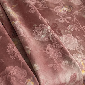 Può includere: Un tessuto rosa polvere caratterizzato da un motivo floreale ripetuto di rose in fiore e foglie. Il materiale presenta una consistenza morbida simile al velluto con sottili variazioni di colore, inclusi tocchi di giallo e crema nei disegni.