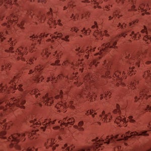 May include: A close-up of a textured, reddish-brown fabric with a repeating pattern of dark brown roses and leaves. The fabric has a slightly wrinkled appearance, suggesting it is draped or folded. The floral design is subtly embossed.