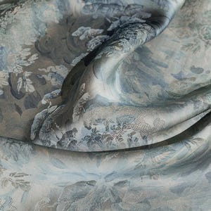 Può includere: Primo piano di un tessuto fluido con un motivo floreale e paesaggistico dettagliato nelle tonalità del blu, grigio e bianco. Il tessuto ha una lucentezza sottile, con dettagli intricati nel design. Le pieghe e i drappeggi del tessuto creano profondità e consistenza.