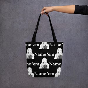 Puede incluir: Bolso tote negro con un patrón repetido de un retrato de mujer y la frase "Name 'em" en blanco. El bolso tiene asas negras y está sostenido por una mano.