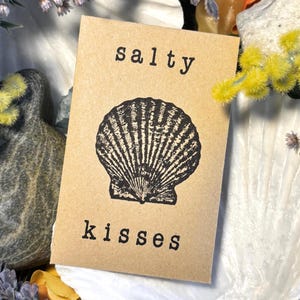 May include: A kraft paper card with the words "salty kisses" and a black seashell illustration. The card is surrounded by dried flowers and a large white seashell.