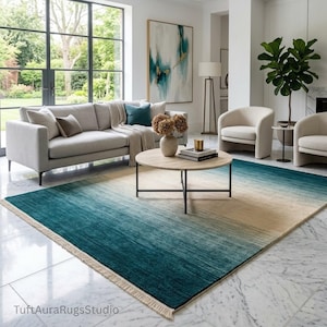 May include: A rectangular rug with a teal to beige gradient design, placed in a living room. The rug features a fringed edge and complements the neutral tones of the sofa and chairs. The rug's dimensions are suitable for a modern living space.