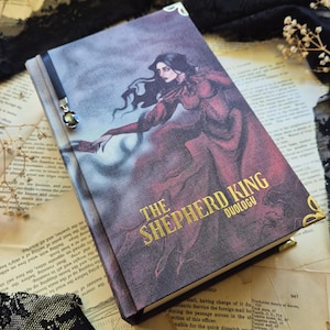 Op de afbeelding: Een hardcover boek getiteld The Shepherd King Duology met een donkerpaarse en rode omslagillustratie van een persoon in een zwierige jurk. Het boek is voorzien van goudkleurige metalen hoekbeschermers en een zwart leeslint met een kristallen bedel.