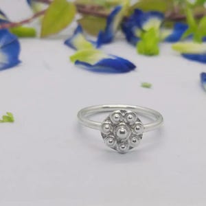 May include: A silver ring with a floral design. The ring has a circular cluster of small, round beads at the centre. It is set against a white background with blue and green flower petals scattered around.