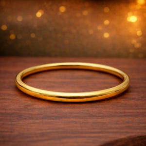Może przedstawiać: Polerowana bransoletka typu bangle w kolorze złotym, o gładkiej, zaokrąglonej powierzchni i kolistym kształcie. Biżuteria posiada ciągły, bezszwowy design z odblaskowym metalicznym wykończeniem, które równomiernie odbija światło.
