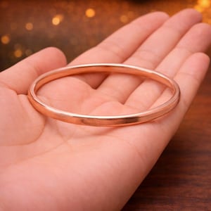 Może przedstawiać: Solidna, okrągła bransoletka typu bangle z gładkim, polerowanym wykończeniem w kolorze różowego złota. Metalowa obręcz ma spójny, zaokrąglony profil i odblaskową powierzchnię, która chwyta światło, tworząc prostą, ciągłą pętlę na nadgarstek.