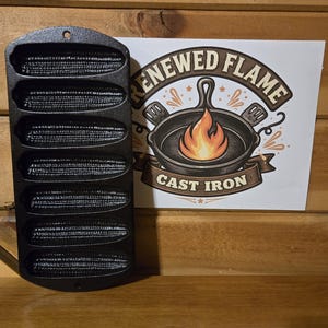 May include: Black cast iron corn cob pan with six individual molds. A white sign displays a graphic of a cast iron skillet with a flame and the words "Renewed Flame Cast Iron". The pan is on a wooden surface.