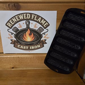 May include: A black cast iron cornbread pan with six corn-shaped molds. A white sign displays the text "Renewed Flame Cast Iron" with a skillet and flames graphic. The pan is on a wooden surface.