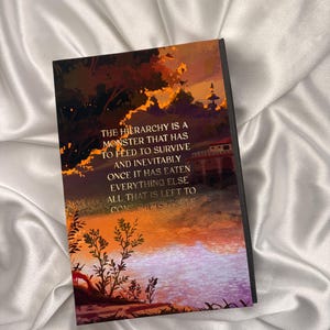 May include: A hardcover book with a vibrant illustration of a sunset over a body of water. The cover features text that reads, "THE HIERARCHY IS A MONSTER THAT HAS TO FEED TO SURVIVE AND INEVITABLY ONCE IT HAS EATEN EVERYTHING ELSE ALL THAT IS LEFT TO CONSUME IS ITSELF."