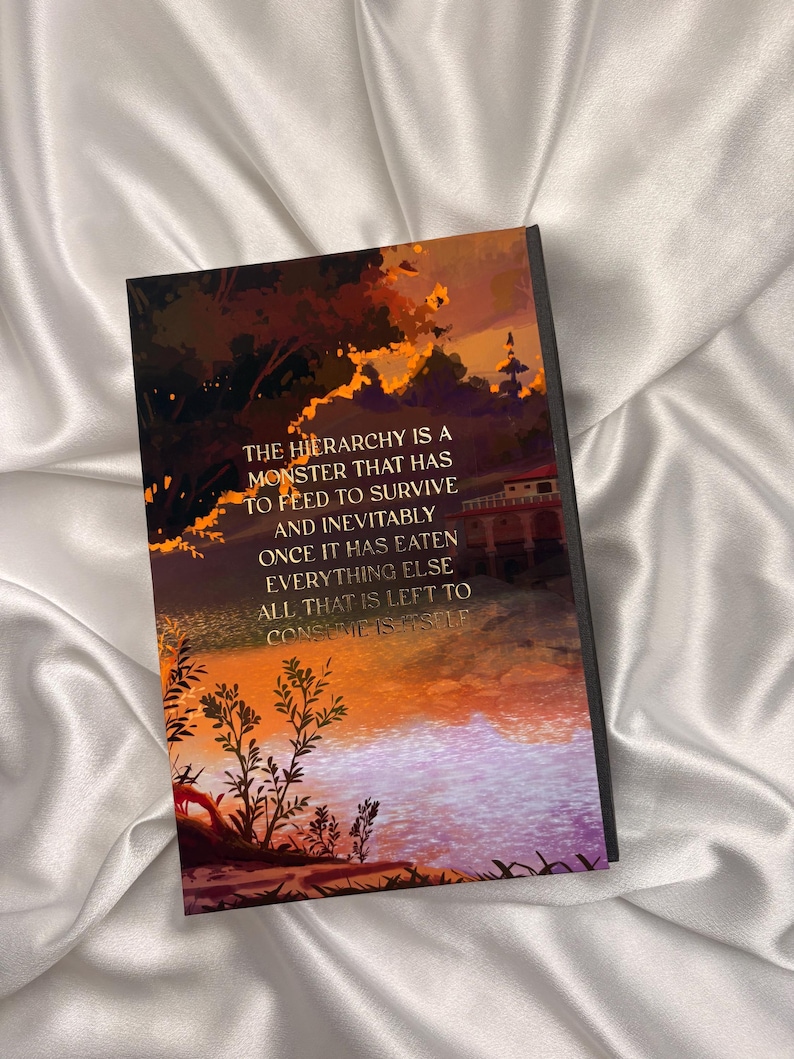 May include: A hardcover book with a vibrant illustration of a sunset over a body of water. The cover features text that reads, "THE HIERARCHY IS A MONSTER THAT HAS TO FEED TO SURVIVE AND INEVITABLY ONCE IT HAS EATEN EVERYTHING ELSE ALL THAT IS LEFT TO CONSUME IS ITSELF."