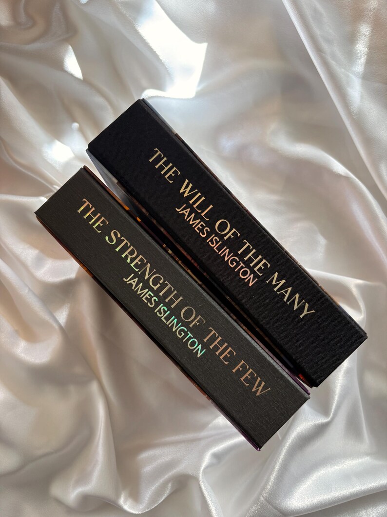 May include: Two hardcover books with black covers and gold lettering. The top book reads "THE WILL OF THE MANY" and "JAMES ISLINGTON." The bottom book reads "THE STRENGTH OF THE FEW" and "JAMES ISLINGTON."