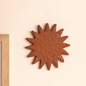 May include: A terracotta-coloured sun wall decoration with a textured, hammered surface. The circular centre features radiating triangular points. Small, hand-painted gold dots are arranged in triangular patterns near the tips of several sun rays.