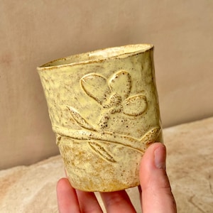 May include: A light yellow ceramic cup with a speckled texture and a raised relief design of a single flower on a stem. The cup has a cylindrical shape with a slightly flared rim and a mottled finish featuring darker brown spots across the surface.