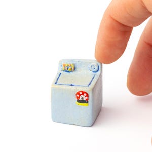 May include: A miniature light blue washing machine figurine with a red and white mushroom design on the front. The top has a small control panel with yellow, blue, and orange details. The figurine is held by a finger.