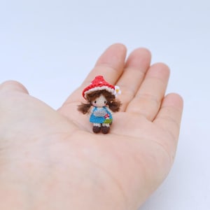 以下が含まれることがあります： 茶色のツインテールの髪をしたミニチュアの編みぐるみ人形。白い水玉模様の赤いキノコ型の帽子、水色のドレス、ダークブラウンの靴を着用しています。帽子の横には小さな白い花の飾りが付いています。