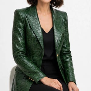 Peut inclure: Un blazer vert foncé avec un motif texturé en relief façon crocodile, doté d'un revers cranté, de manches longues et de boutons dorés sur les poignets et la fermeture avant. Le blazer est porté sur un haut noir et un pantalon noir, avec un sac à main noir.
