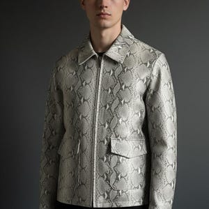 Puede incluir: Chaqueta con estampado de piel de serpiente en gris claro y blanco, con cierre de cremallera y dos bolsillos delanteros. La chaqueta tiene un cuello clásico y se lleva sobre una camisa negra. El fondo es gris liso.
