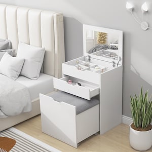 Puede incluir: Un tocador blanco con espejo abatible, cajones y un taburete extraíble. El tocador tiene compartimentos para guardar joyas y cosméticos. El taburete tiene un asiento tapizado gris. El tocador está junto a una cama con almohadas blancas.