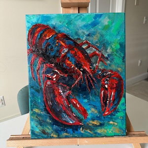 Op de afbeelding: Origineel acrylverfschilderij van een kreeft. Het kunstwerk toont een levendige rode kreeft met blauwe accenten tegen een turquoise achtergrond. De kreeft is geschilderd met gedurfde penseelstreken, waardoor een textuur en dynamisch effect ontstaat. Het schilderij wordt tentoongesteld op een houten ezel.